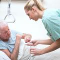 Ein älterer Mann liegt in einem Krankenbett und erhält von einer medizinischen Fachkraft Tabletten und ein Glas Wasser. 