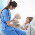 Eine Krankenpflegeassistentin betreut ein kleines Mädchen, welches in einem Krankenbett liegt. Die Krankenpflegeassistentin sitzt auf dem Krankenbett des Mädchens und hält ihr aufmunternd ein Kuscheltier vor das Gesicht. 