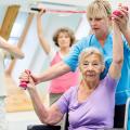 Eine Ergotherapeutin betreut eine ältere Dame beim Sporttreiben. Im Hintergrund befinden sich ältere Personen, die ebenfalls sportliche Aktivitäten ausüben. 