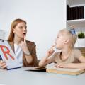 Eine Logopädin übt mit einem Kind die Aussprache des Buchstabens "R". Der Buchstabe steht auf einem Blatt Papier und wird dem Kind deutlich gezeigt. Vor dem Kind befindet sich ein Buch auf einem Schreibtisch. 