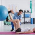 Ein Physiotherapeut betreut eine Patientin in einem Raum eines Physiozentrums. Die Frau liegt mit dem Rücken auf dem Boden und drückt mit ihren Beinen einen Gymnastikball an eine Wand. Der Therapeut kniet dabei neben ihr und stützt ihren Fuß. 