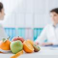 Im Fokus stehen verschiedene Obstsorten und ein Maßband. Im Hintergrund sitzen sich zwei Ärztinnen gegenüber. 