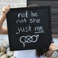 Eine Person hält eine Tafel in den Händen mit der Aufschrift "not he not she just me" (zu Deutsch: nicht er nicht sie nur ich). Darunter sind die Symbole für Männlich und Weiblich durch das Zeichen für Unendlichkeit miteinander verbunden. 