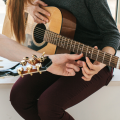 Abgebildet sind zwei Personen, die jeweils eine Gitarre in den Händen halten. Die Person links macht sich zusätzlich Notizen auf einem Blatt Papier. 