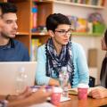 Vier Personen sitzen und unterhalten sich an einem Tisch. Auf dem Tisch liegen Schreibutensilien, Bücher und ein Laptop.