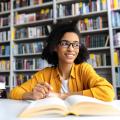 Eine Frau sitzt an einem Tisch in einer Bibliothek. Vor ihr befindet sich ein geöffnetes Buch und in ihrer Hand hält sie einen Stift.