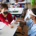 Eine Krankenschwester misst die Temperatur eines Schuljungen in einem Klassenraum. Beide tragen einen Mund- und Nasenschutz. 