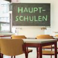 Abgebildet ist ein Klassenraum. Auf der Tafel im Klassenraum steht das Wort "Hauptschulen".