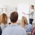 Mehrere Personen sitzen in einem Schulungsraum und schauen nach vorne, wo eine Frau etwas an einem Flipchart erklärt. 