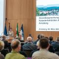 Abgebildet sind mehrere Personen, die sich in einem Sitzungssaal befinden. Eine Rednerin steht an einem Pult und andere Personen sitzen ihr gegenüber. 