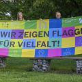 Vier Mitarbeiterinnen und Mitarbeiter der Bezirksregierung Arnsberg halten eine Flagge hoch. Die Flagge beinhaltet viele unterschiedliche Farben. Es steht geschrieben: "Wir zeigen Flagge für Vielfalt!".