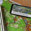 Abgebildet ist ein Handy, auf dessen Bildschirm "Änderungen des Regionalplanes Arnsberg – Teilabschnitt Kreis Soest und Hochsauerlandkreis" steht. Unter dem Handy befindet sich eine topographische Karte. 