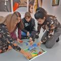 Vier Studentinnen knien auf dem Boden in einem Museumssaal. Sie legen farbige Bildelemente zu einer Collage zusammen. An den Wänden im Hintergrund Ausschnitte von modernen Kunstwerken. 