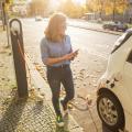Junge Frau steht neben dem Elektroauto und schaut auf ihr Smartphone. Das Mietauto wird an der Ladestation für Elektrofahrzeuge aufgeladen.