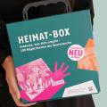 Abgebildet ist eine Person, die eine Box mit der Aufschrift "Heimat-Box" in den Händen hält.